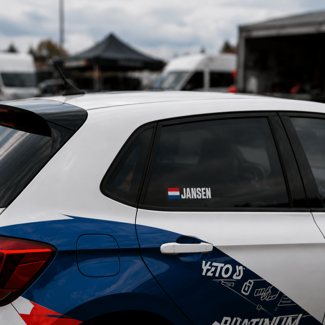 Rally naamsticker met vlag op de zijruit van een rallyauto in een paddockomgeving